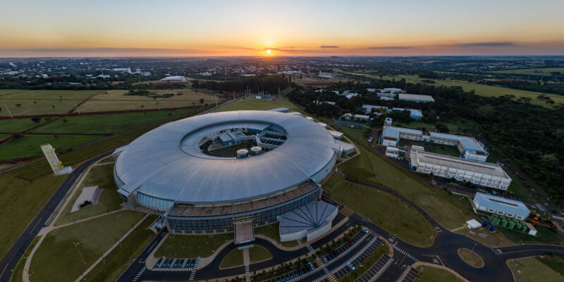Fotografia aérea ao entardecer do edifício do Sirius, acelerador de partículas do CNPEM, em Campinas (SP). A estrutura tem formato circular, com cobertura metálica em tons de prata e pátio central aberto. Ao redor, campus com outros prédios institucionais, áreas verdes e estacionamento. Ao fundo, a cidade e o sol no horizonte sob céu alaranjado.