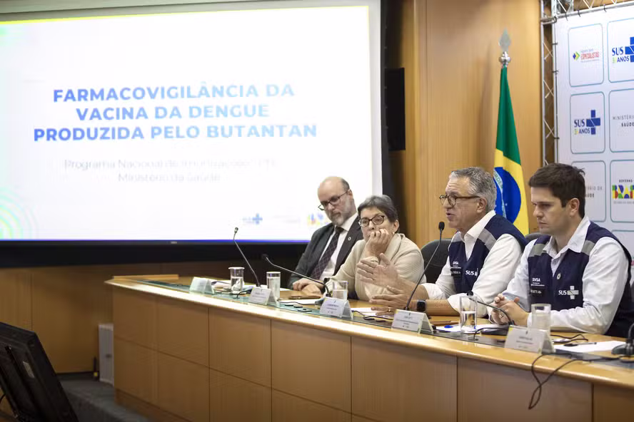 Mesa de coletiva de imprensa com três homens ao fundo de bandeiras do Brasil e do SUS; tela ao centro exibe os dizeres 'Farmacovigilância da vacina da dengue produzida pelo Butantan'