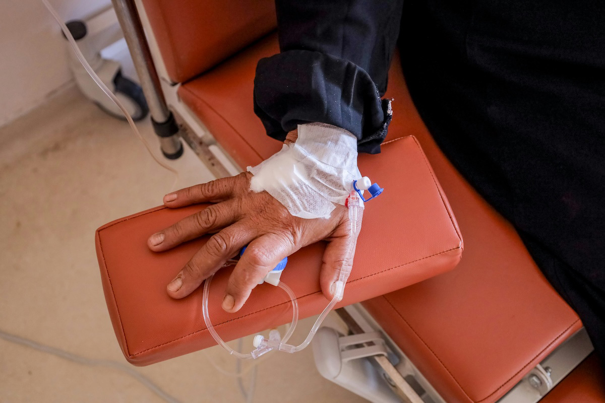 Close-up photograph of a hand resting on the arm of an orange synthetic-leather armchair in a hospital setting. The wrist is bandaged with white gauze and connected to an intravenous catheter with a transparent tube. The person is wearing a long dark-sleeved top. An off-white floor and part of an IV stand are visible in the background.