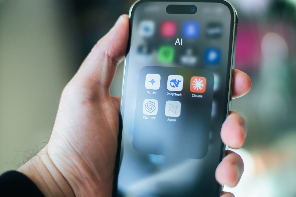 Close-up photograph of a hand holding a smartphone, with the screen showing an app folder labeled "AI" containing the icons for Gemini, DeepSeek, Claude, ChatGPT, and Auren. The background is an out-of-focus indoor setting in shades of pink and green.