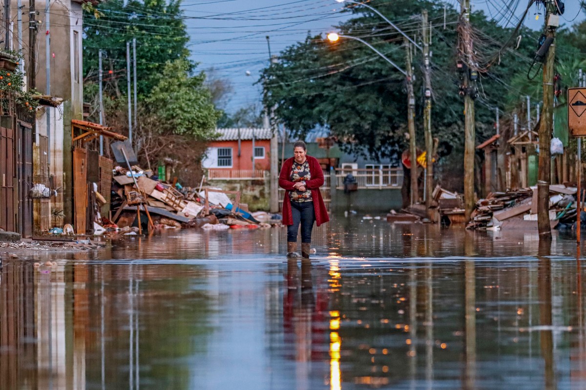 Mulher caminha por rua alagada, cercada por casas, postes e entulhos, após uma enchente em área urbana. A imagem evidencia os impactos de inundações sobre comunidades, infraestrutura e rotina da população