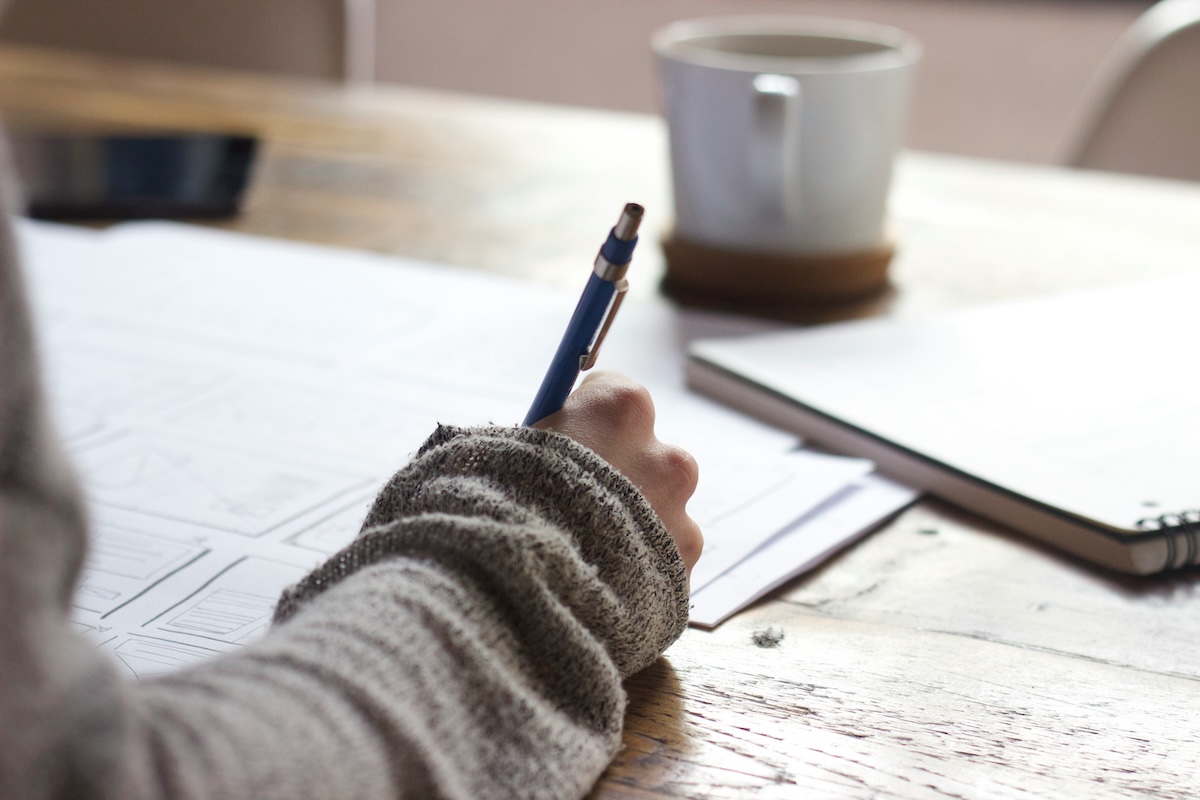 Close-up photograph showing an arm clad in a gray knit sweater, holding a blue pen over a notebook on a wooden table. In the background, a white coffee mug sits on a wooden coaster beside a spiral-bound notebook.