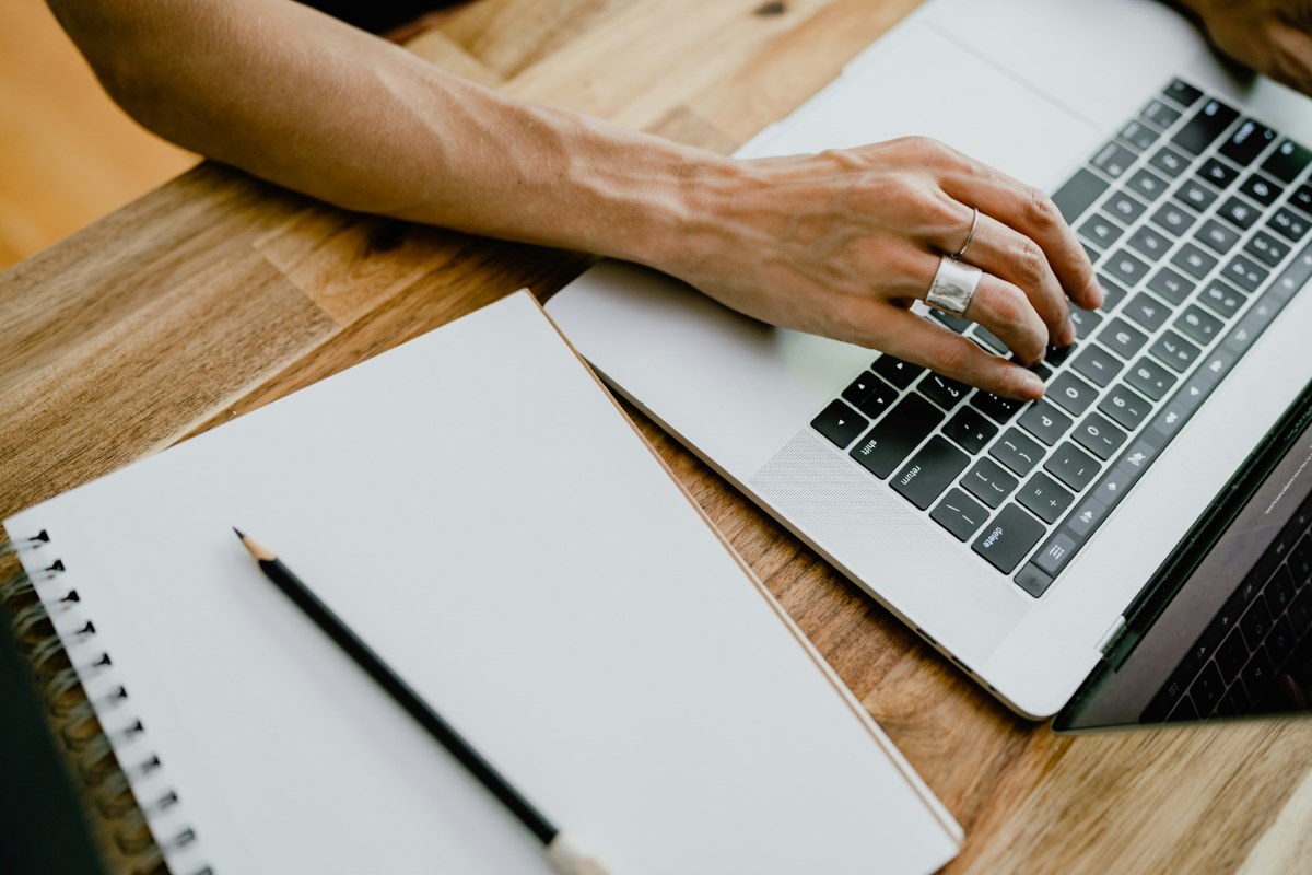 Pessoa digita em um notebook ao lado de um caderno em branco e um lápis, sobre uma mesa de madeira. A cena remete ao processo de escrita, organização de ideias e uso de ferramentas digitais