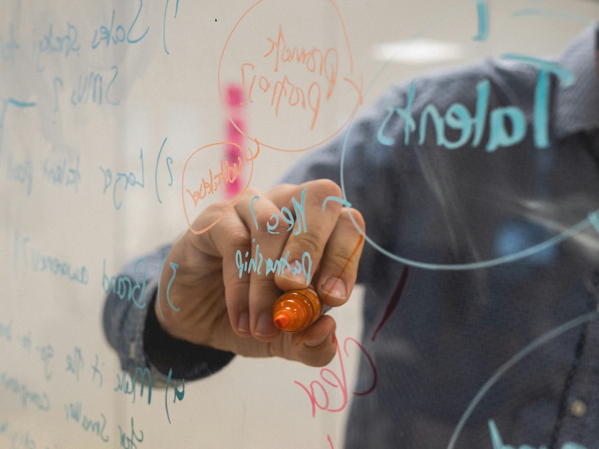 Close-up photograph of a person clad in a blue shirt sleeve writing with an orange marker on a transparent glass surface. In the background, blue, orange, and pink notes connect terms such as "Talent," "Growth," and "Leadership" with circles and arrows, evoking a corporate brainstorming session.