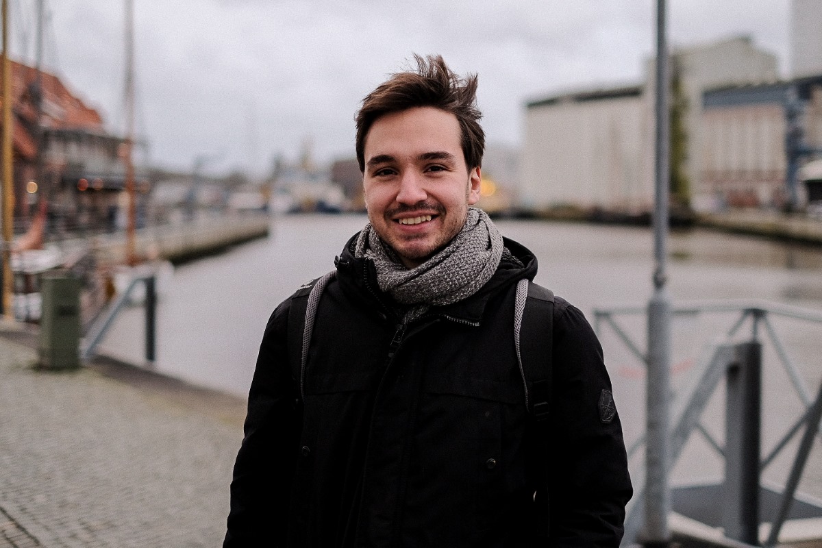 Portrait of Lennart Banse outdoors in an urban area, with a canal and buildings out of focus in the background. The researcher is smiling, dressed in a coat and scarf in what appears to be a cold setting.