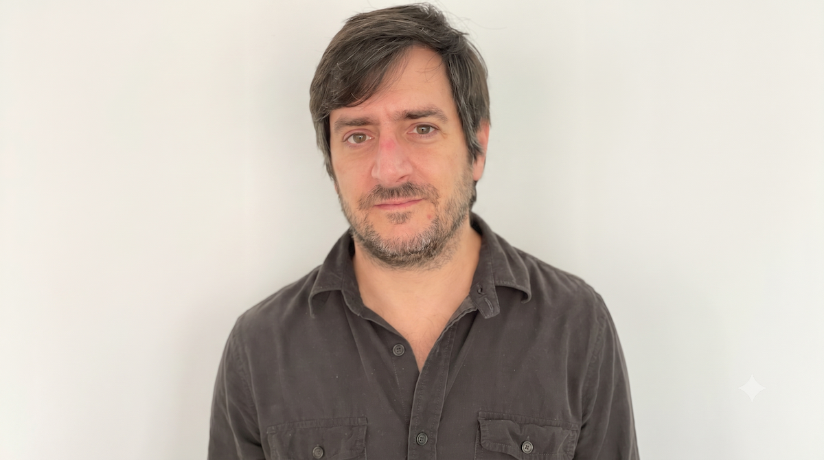 Head-and-shoulders portrait, against a light gray background, of a man with brown hair and a short, sparse beard, wearing a dark shirt and looking directly at the camera with a neutral expression.