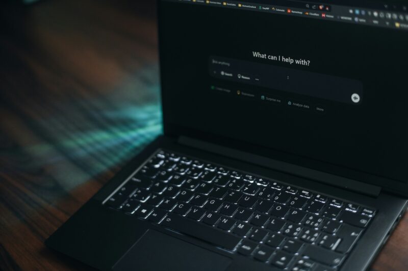 Tela de notebook em ambiente escuro exibe interface de chatbot de inteligência artificial com a pergunta "What can I help with?" e opções de busca, raciocínio e criação de imagem. Teclado retroiluminado em primeiro plano, sobre mesa de madeira com reflexos de luz azulada
