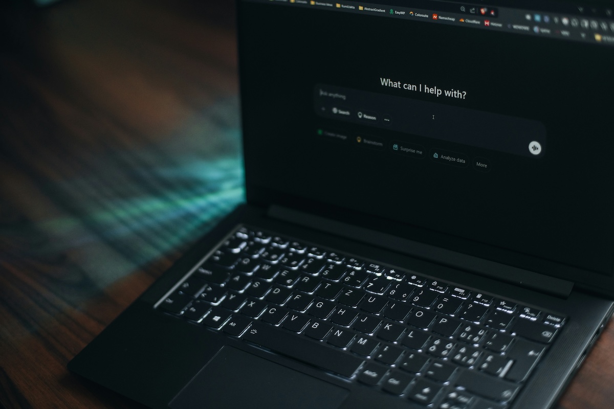 An open laptop in a dark environment, with an AI chatbot interface on the screen displaying the question "What can I help with?" and options to search, reason, or create an image. The laptop, with a backlit keyboard, is on a wooden desk illuminated by streaks of blue light.