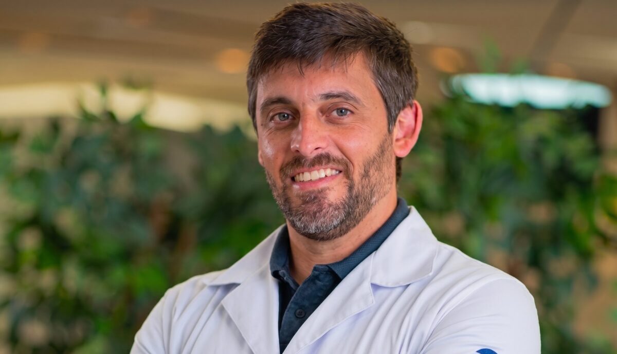 A white man with short brown hair and a neatly trimmed short beard smiles with his arms crossed. He is wearing a white lab coat over a dark polo shirt, with embroidered lettering on the lab coat pocket. The background is an indoor setting with blurred green plants.