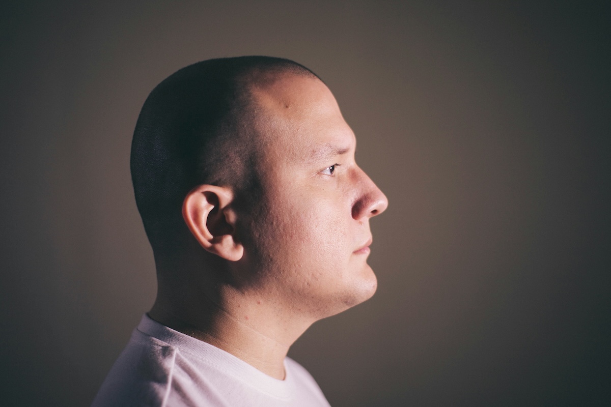 Retrato de perfil de um homem com a cabeça raspada, olhando para a direita. A iluminação lateral destaca o contorno do rosto, orelha e mandíbula contra um fundo escuro e desfocado. Ele veste uma camiseta branca.
