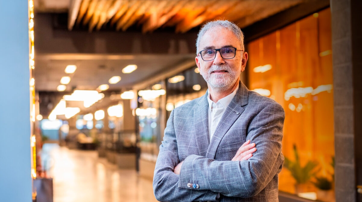 A white man with gray hair and beard, wearing dark-rimmed glasses, smiles slightly and stands with his arms crossed. He is wearing a gray plaid blazer over a white collared shirt. The photograph is set in a wide interior corridor with pale flooring, warm lighting, and out-of-focus display windows in the background.