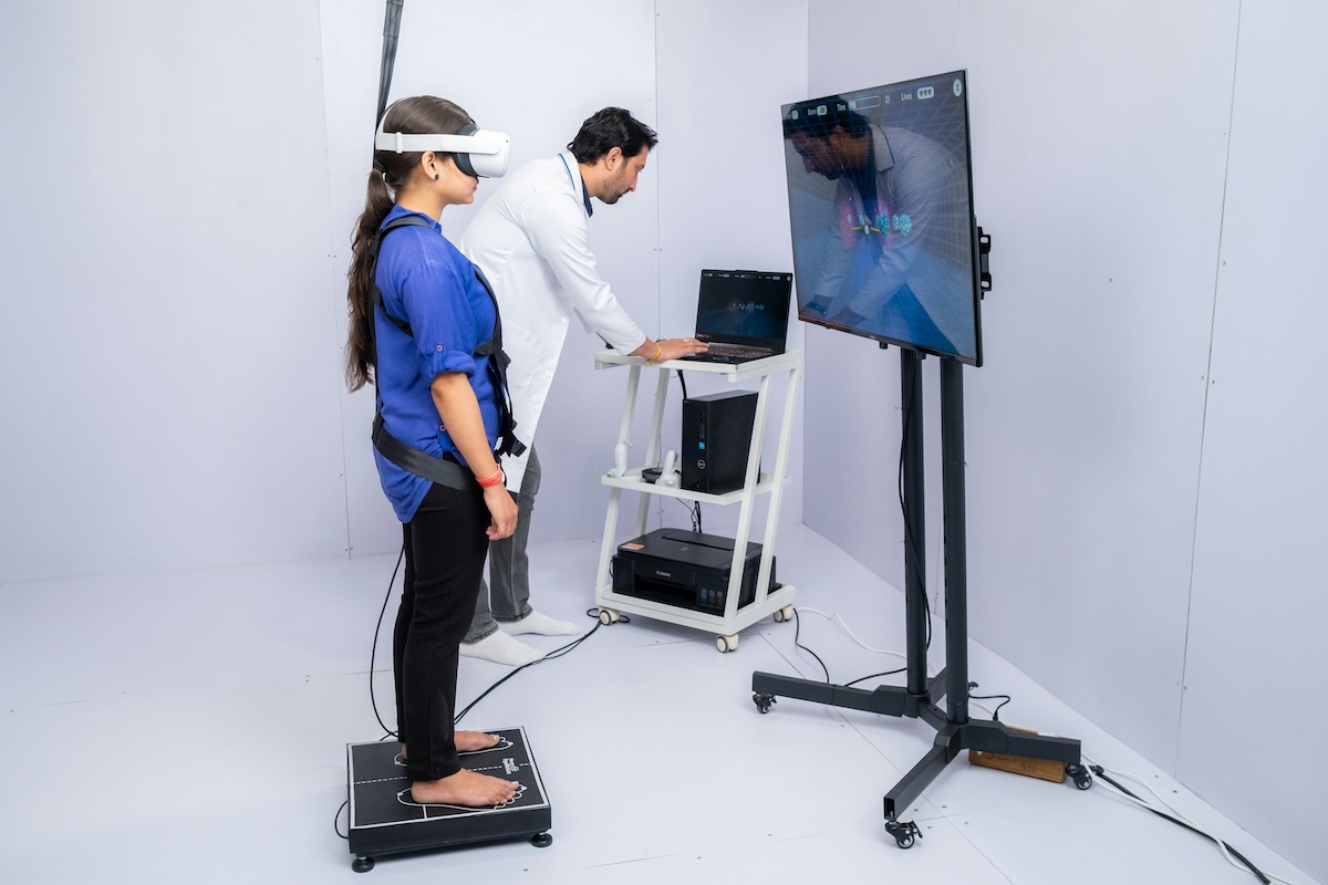 Mulher de cabelo escuro preso em rabo de cavalo, descalça, vestindo blusa azul e calça preta, usa óculos de realidade virtual e colete com sensores presa a uma plataforma de equilíbrio; ao lado, homem de jaleco branco opera um notebook em um carrinho com computador e impressora, enquanto uma tela grande à frente exibe a captura de movimento em tempo real.